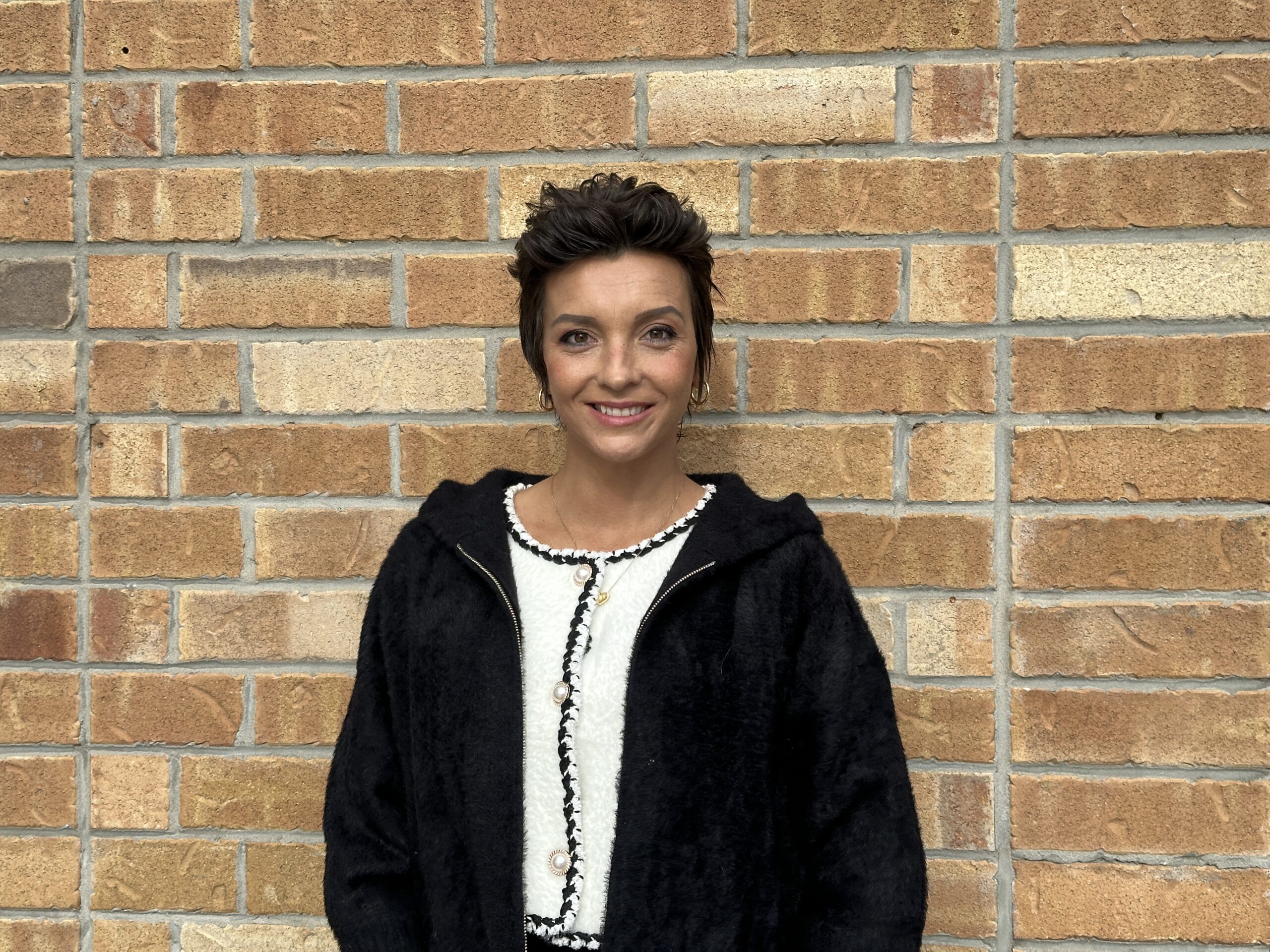 Portrait of Joanna, a mother of three facing stage three breast cancer, standing in front of a brick wall with a black sweater during the holiday season.